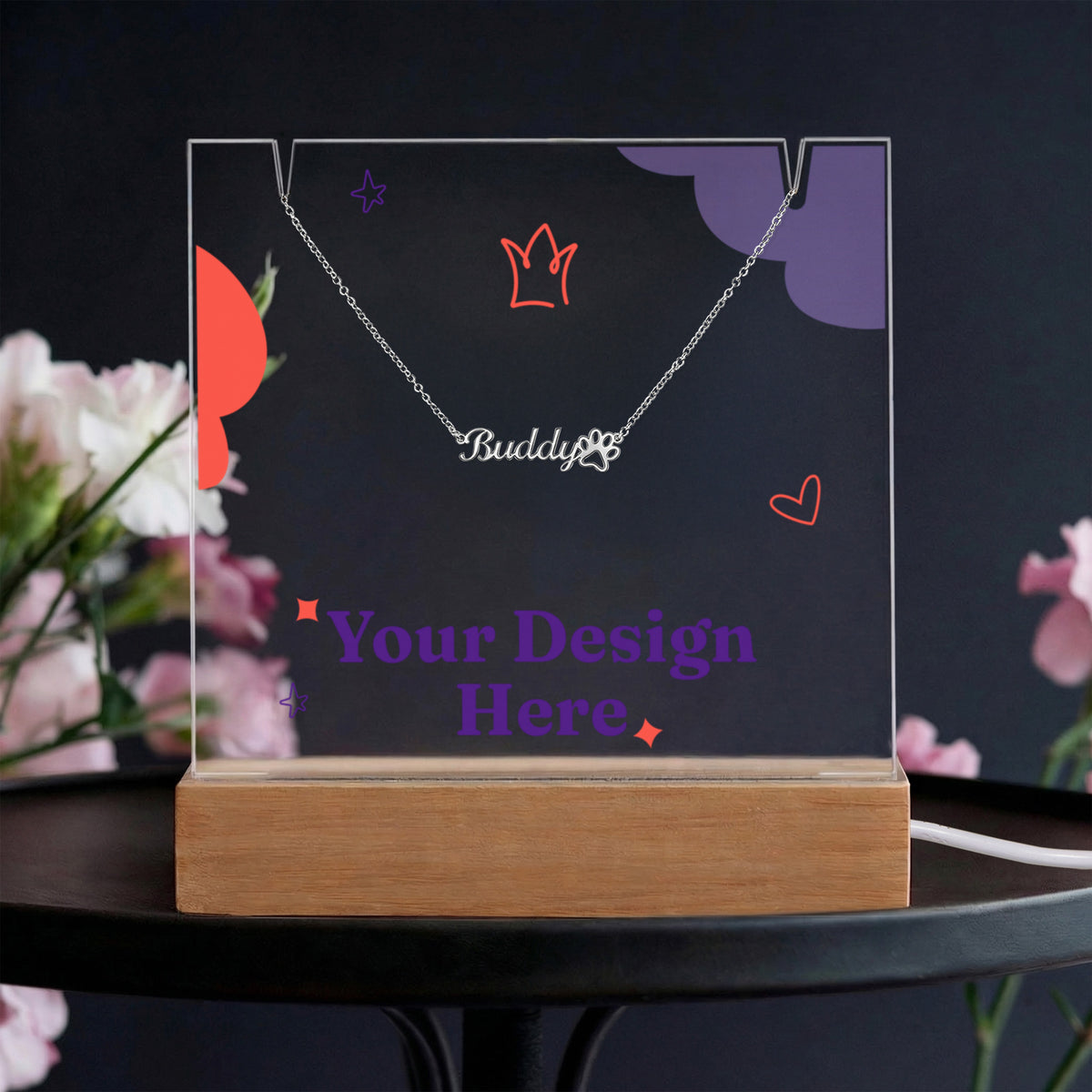 Acrylic keepsake and paw print name necklace on a dark table beside pink flowers | Pet Photo Keepsake Light and Paw Necklace