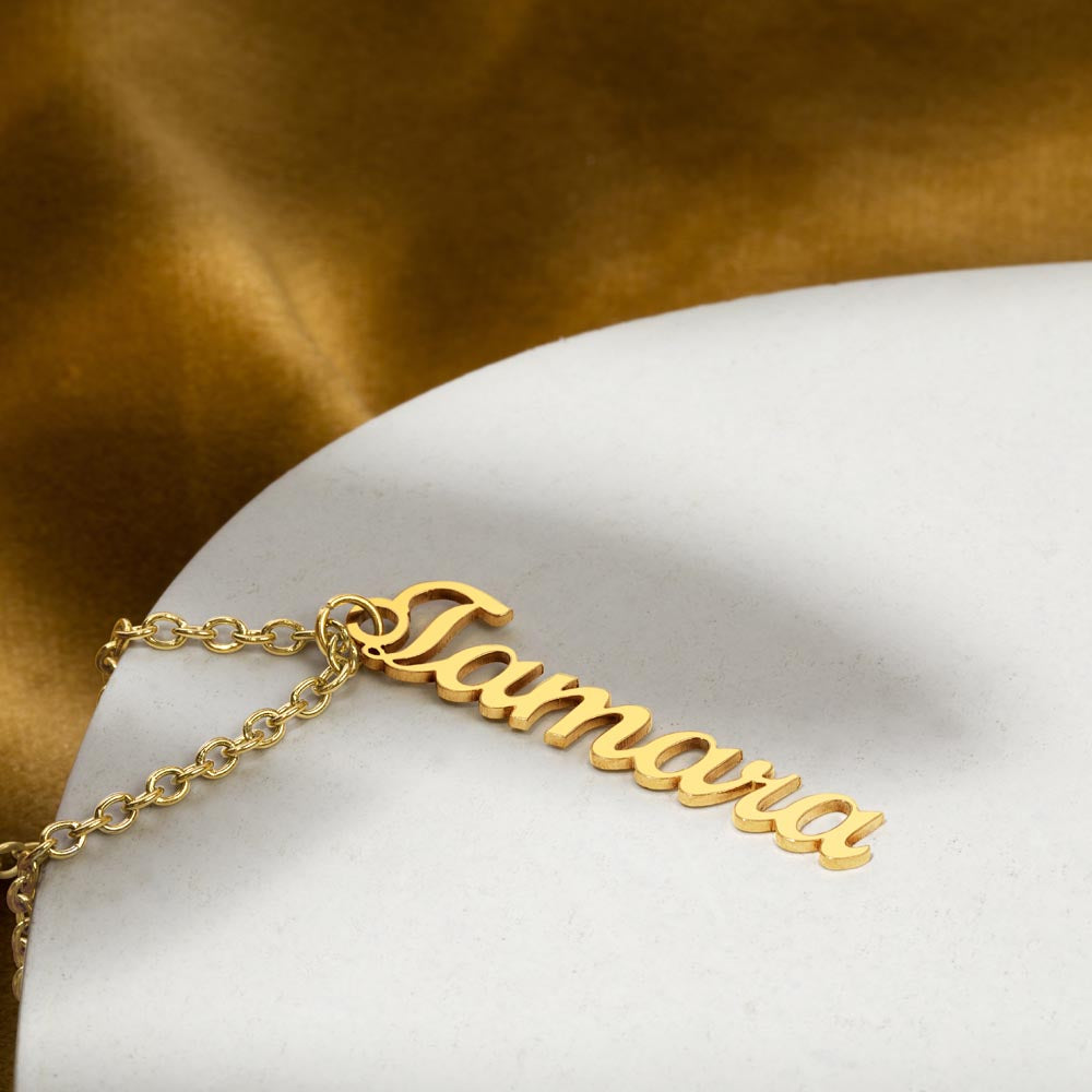 Gold finish name pendant lying on a gold and white surface | Vertical Name Necklace