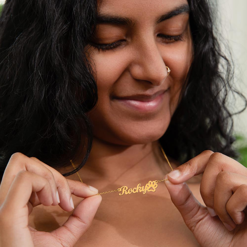 Smiling woman holding the gold finish paw print name necklace at her neckline | Dog Mom Paw Print Name Necklace with Message Card
