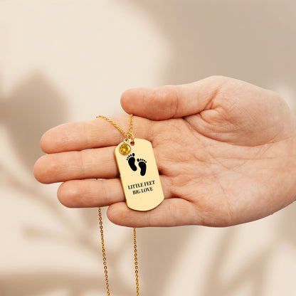 Tag pendant engraved with baby footprints and LITTLE FEET BIG LOVE with a birthstone charm, held in a hand